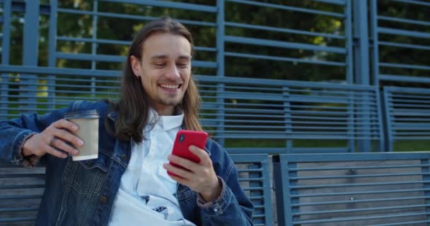 Joven alegre con el pelo largo sosteniendo la taza de café de papel mientras mira el vídeo divertido. Millennial chico con pendientes usando teléfono inteligente y riendo mientras está sentado en el banco al aire libre. — Vídeo de stock