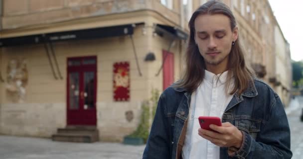 Ung långhårig kille tittar på telefonskärmen och skrattar samtidigt få roligt budskap. Skägg tusenårig man skriva när du använder sin smartphone och promenader på stadens gata. — Stockvideo