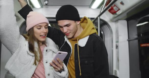 Millennial usmívající se pár dívá na obrazovku telefonu při sledování videoklipu. Mladý kluk a jeho přítelkyně sdílení sluchátka při poslechu hudby a jít do veřejné dopravy. — Stock video