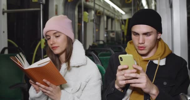 Jeune beau mec utilisant son smartphone tout en étant assis près de sa petite amie. Millennial attrayant livre de lecture fille tout en allant dans les transports en commun avec son petit ami. Concept de vie réelle. — Video