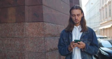 Genç adam akıllı telefon kullanıyor ve şehir caddesinde yürürken başını kaldırıyor. Sosyal medyada sohbet ederken uzun saçlı, mesaj yazan milenyum adamı. Gerçek hayat kavramı.