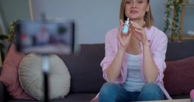 Genç bir kadın evdeki kanepede otururken akıllı telefon kamerasıyla video kaydediyor. Kozmetik üzerinde inceleme yaparken kadın vloger konuşuyor ve ürünü tutuyor. Güzellik vloglama kavramı.
