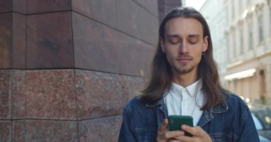 Dışarıda yürürken cep telefonu kullanan genç adamın yakın görüntüsü. Uzun saçlı yakışıklı adam. Şehir sokağındaki telefon ekranına bakıyor. Gerçek hayat kavramı.