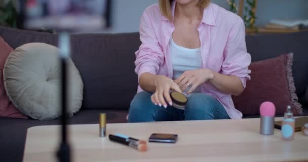 Glimlachend meisje praten en tonen aan de camera cosmetische product tijdens het zitten op de bank thuis.Vrolijke vloger opname op smartphone make-up review. Begrip schoonheid en vlogging. — Stockvideo