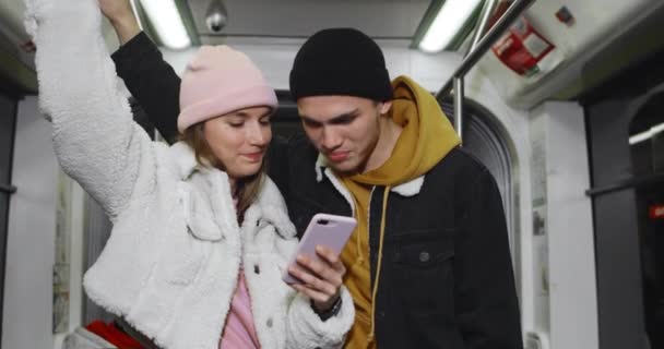 Веселая пара тысячелетия смотрит на смартфон во время поездки в метро. Молодая красивая девушка держит телефон, показывая экран своему парню. Концепция отношений. — стоковое видео