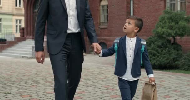 Beskär utsikt över manlig person i kostym håller hand i hand med liten pojke när du går på gatan. Barn med ryggsäck tittar och pratar med far när du går till skolan på morgonen. — Stockvideo