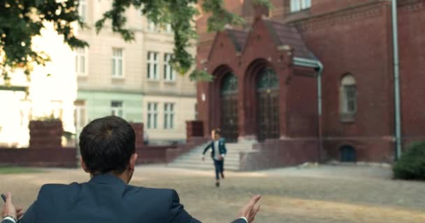 Baksidan av mannen i kostym väntar på sitt barn efter lektioner nära skolan byggnad. Glada pojke med väska klädd i uniform som springer i far omfamna. Begreppet familj. — Stockvideo