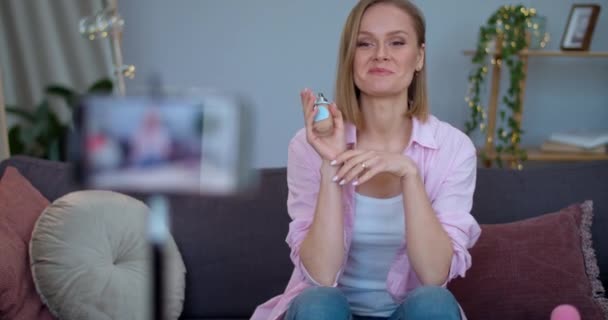 Blondýny žena vloger dělat video recenzi o make-up produkt, zatímco sedí na gauči doma. Veselá dívka mluví a ukazuje základy kůže při nahrávání sebe na smartphone. — Stock video