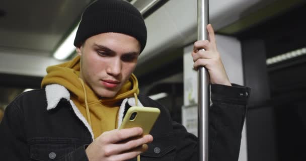 Młody, przystojny student korzystający ze smartfona i uśmiechnięty podczas dojazdu środkami transportu publicznego. Millennial facet patrząc na ekran telefonu podczas pisania wiadomości w mediach społecznościowych. — Wideo stockowe