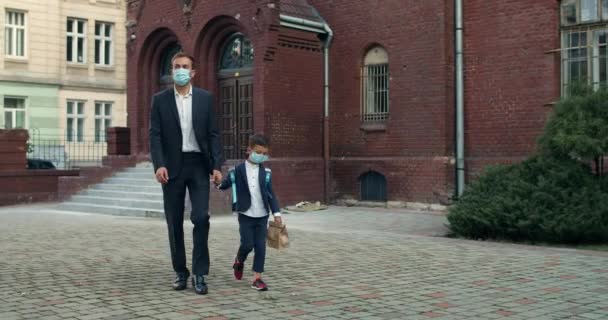 Geschäftsmann im Anzug umarmt seinen Jungen, während er zur Schule geht. Sohn mit Tasche und sein Vater, der beim Gehen medizinische Masken trägt und Hand in Hand geht.. — Stockvideo