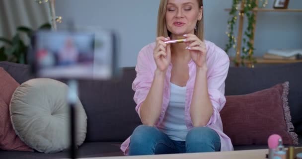Atractiva vloger femenina haciendo revisión de vídeo sobre el producto de maquillaje mientras está sentado en el sofá. Mujer joven hablando y mostrando rímel mientras se graba en el teléfono inteligente para vlog. — Vídeos de Stock