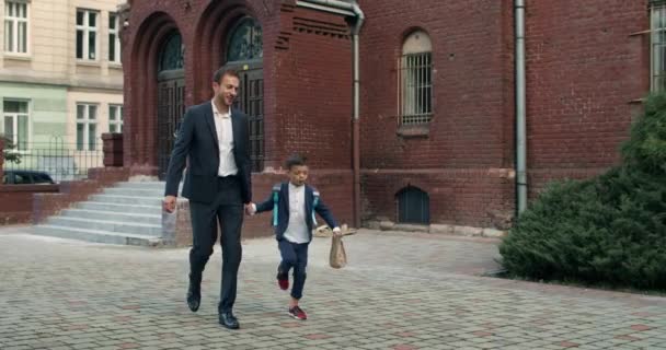 Liten pojke med väska hoppar hapilly medan du går och håller hand i sin leende far. Glad man i kostym som tar sin son till skolan. Begreppet barn och utbildning. — Stockvideo