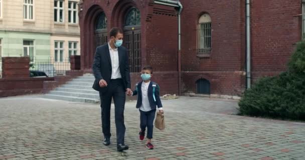 Μικρό παιδί με σακίδιο και ο πατέρας του με ιατρικές μάσκες κρατώντας το χέρι στο χέρι ενώ περπατούσε. Επιχειρηματίας με κοστούμι πηγαίνει το παιδί του στο σχολείο. Έννοια των παιδιών και εκπαίδευση. — Αρχείο Βίντεο