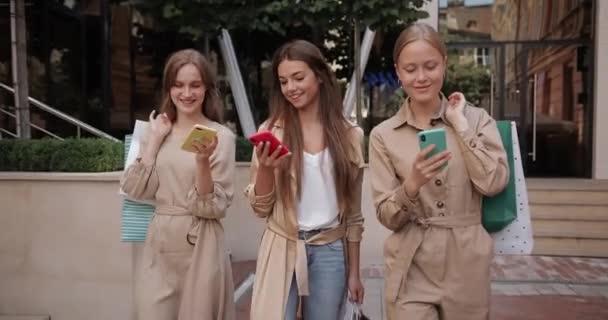 Закройте глаза подругам, использующим свои смартфоны во время прогулки по городской улице. Привлекательные молодые женщины с бумажными пакетами смеются, глядя на экраны телефонов после покупок. — стоковое видео