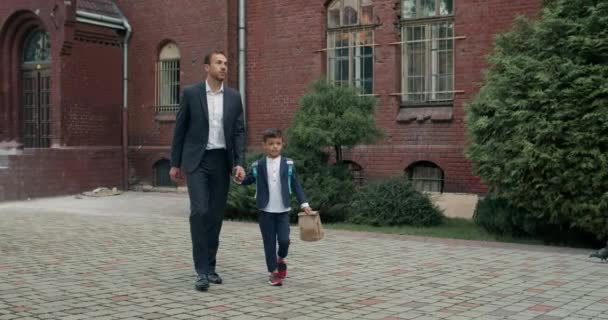 Guapo padre hombre de negocios y niño pequeño con bolsa hablando mientras toma de la mano. Hombre con su lindo hijo caminando en el patio de la escuela. Concepto de educación y niños. — Vídeo de stock