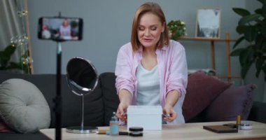 Neşeli kadın vlogger kaydedicisi evde kanepede otururken akıllı telefondan paketleri açıyor. Güzel kız sosyal ağ etkileyicisi kutuyu açıyor ve sürpriz yüz ifadesi takınıyor.