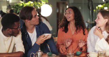 Gözlüklü güzel bir kadının hoş bir kafede arkadaşlarıyla komik bir hikaye anlatırken el kol hareketi yaptığı görüntüler. Neşeli genç erkekler ve kızlar iletişim kuruyor ve gülüyor. Duygular kavramı..