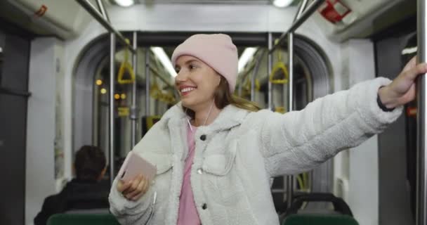 Vista colheita de alegre jovem mulher vestindo roupas elegantes usando seu smartphone. Millennial atraente estudante em fones de ouvido sorrindo enquanto vai no transporte público. — Vídeo de Stock