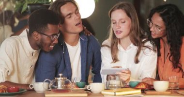 Güzel genç bir kadın, arkadaşlarına video gösterirken akıllı telefonu yatay olarak tutuyor. Çok ırklı bir grup öğrenci birlikte kafede vakit geçirirken gülümseyip telefon ekranına bakıyorlar..
