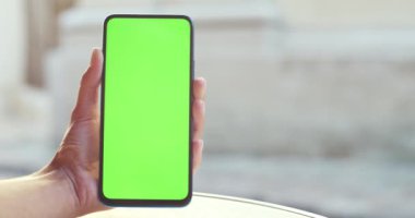 Akıllı telefonu tutan ve model şablon ekranına basan kadının yakın görüntüsü. Yeşil ekran ve krom anahtar kavramı. Arka plandan geçen insanlar.