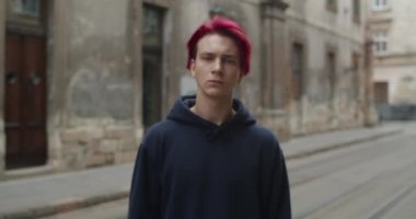 Kırmızıya boyanmış saçlı ve kameraya bakan depresif adamın görüntüsü. Bin yıllık hippi portresi. Küpeli, siyah kapüşonlu bir adam. Boş şehir caddesinde duruyor..