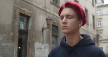 Küpeli, koyu renk kapüşonlu, dışarıda yürüyen üzgün genç adamın yakın görüntüsü. Kırmızıya boyanmış saçlı hippi milenyum çocuğu eski şehir sokağında depresif görünüyor. Yaşam tarzı kavramı.