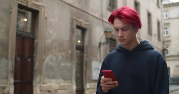 Προβολή των καλλιεργειών χιλιετή hipster τύπος με σκουλαρίκι αναζητούν και κύλιση οθόνη του τηλεφώνου. Νεαρός με βαμμένα κόκκινα μαλλιά χρησιμοποιώντας smartphone ενώ περπατούσε στο παλιό δρόμο της πόλης. Έννοια του τρόπου ζωής. — Αρχείο Βίντεο