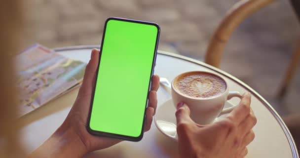Widok na kobietę trzymającą zielony ekran telefonu komórkowego i filiżanki kawy siedząc przy stole na ulicy. Koncepcja klucza chromatycznego i makiety. — Wideo stockowe
