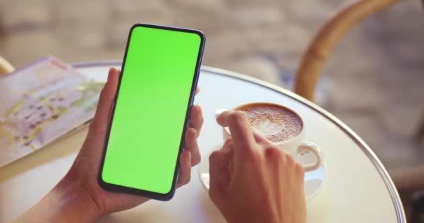 Vista de la cosecha de la persona femenina sosteniendo el teléfono inteligente y desplazándose en la pantalla verde mientras está sentado en la mesa con café y mapa en él. Concepto de maqueta y clave de croma. — Vídeos de Stock