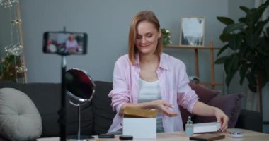 Çekici kadın video kaydedicisi evdeki kanepede otururken akıllı telefondan paketleri açan güzelliği kaydediyor. Etkilenen kız makyaj aksesuarlarıyla birlikte kutuyu açıyor ve sürpriz yüz ifadesi takınıyor..