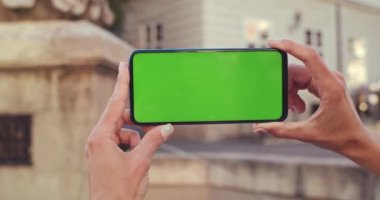 Moda tırnak tasarımlı, akıllı telefonları yatay ekranda tutan kadın elleri yakın çekim görüntüsü. Arka planda eski şehir çeşmesi var. El yapımı ekran ve krom anahtar kavramı.