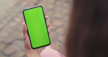 Sokakta otururken akıllı telefonu model ekranla tutan kadının omuz üstü görüntüsü. Krom anahtar ve yeşil ekran kavramı.