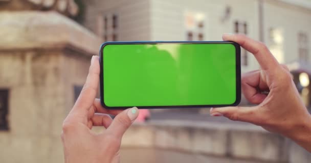 Close up άποψη των γυναικείων χεριών με το σχεδιασμό των νυχιών κρατώντας κινητό τηλέφωνο με πράσινη οθόνη οριζόντια. Παλιά πηγή της πόλης στο παρασκήνιο. Έννοια του mockup και του χρωματικού κλειδιού. — Αρχείο Βίντεο