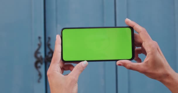 Κλείσιμο προβολής των γυναικείων χεριών κρατώντας το κινητό τηλέφωνο οριζόντια και πιέζοντας στο πράσινο οθόνη πρότυπο. Έννοια του mockup και chroma κλειδί. Μπλε πόρτα στο φόντο. — Αρχείο Βίντεο