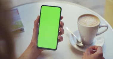 Kahve fincanı ve haritasıyla masada otururken elinde yeşil ekran akıllı telefon tutan kadın gezginin kırpma görüntüsü. Model ve krom anahtar kavramı.