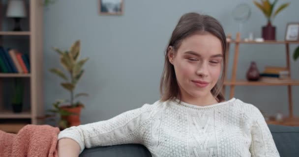 Vue rapprochée de la jeune femme joyeuse souriante tout en regardant la caméra. Portrait d'une femme séduisante posant assise sur un canapé dans une maison confortable. Concept de personnes et d'émotions. — Video