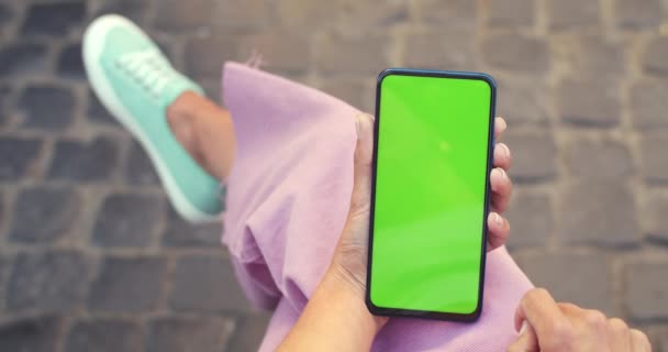 Schnittansicht einer weiblichen Person, die auf dem Bildschirm wischt und drückt, während sie ihr Handy hält und auf der Straße sitzt. Konzept von Chroma Key und Greenscreen. Pflastersteine Hintergrund. — Stockvideo