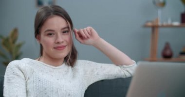 Bin yıllık güzel bir kadının başını çevirip kameraya bakıp gülümseyen dizüstü bilgisayarını kullanmasının portresi. Evdeki kanepede otururken poz veren neşeli kadın görüntüsü.