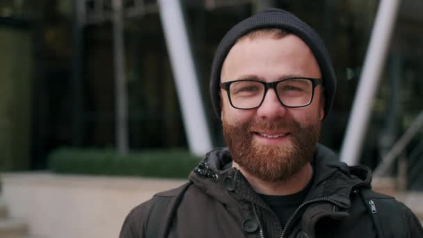 Portrait d'homme barbu joyeux dans les années 30 regardant à la caméra et posant. Vue rapprochée de la belle personne masculine en lunettes portant un chapeau souriant tout en se tenant debout dans la rue. Concept d'emoton. — Video