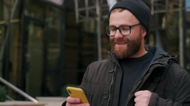 Recorte vista del hombre en gafas mirando a la pantalla del teléfono y sonriendo mientras camina por la calle. Tipo barbudo con bolsa en el hombro desplazándose noticias de redes sociales mientras se utiliza el teléfono inteligente. — Vídeos de Stock