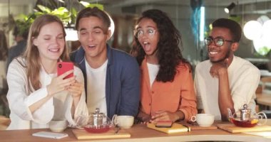 İş arkadaşlarının yemek sırasında iyi vakit geçirirken telefon ekranına bakması. Millennial gülen arkadaşlar modern kafede masada otururken akıllı telefondan fotoğraf izliyorlar..
