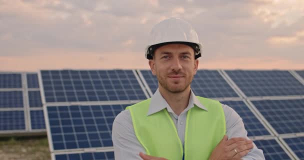 Porträtt av glad manlig ingenjör i skyddande hjälm korsning armar medan du tittar på kameran. Snygg man i uniform som ler när han står på solkraftspark. Begreppet grön energi. — Stockvideo