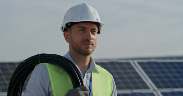 Nahaufnahme eines gut aussehenden männlichen Ingenieurs in Uniform, der an einem Solarkraftwerk spaziert. Kaukasischer Mann mit Harthelm, der Gegenstand untersucht und Kabel trägt. Konzept der grünen Energie.