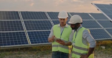 Güneş çiftliğinde duran çeşitli erkek mühendislerin ekin manzarası ve verimli bir inşaat planı tartışması. Koruyucu miğferli ve üniformalı iki adam konuşuyor ve tablet ekranlara bakıyor..