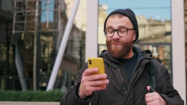 Homme heureux dans des lunettes l'air excité et souriant tout en marchant dans la rue. Barbu gars ayant de bonnes nouvelles et se réjouir tout en utilisant un smartphone. Concept de succès et de victoire. — Video