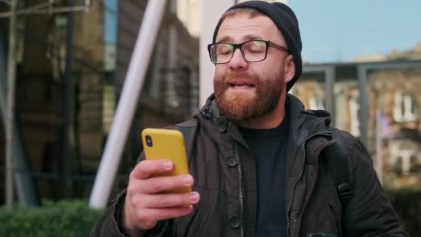 Vue rapprochée de l'homme barbu chantant chanson et drôle de mouvement tout en marchant dans la rue. Gai gars dans les lunettes ayant de bonnes nouvelles et se réjouir tout en utilisant son smartphone. — Video