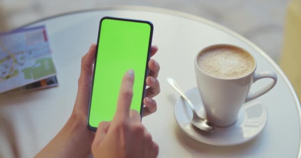 Vista de la cosecha de la persona femenina sosteniendo el teléfono inteligente y presionando en la pantalla de maqueta mientras está sentado en la mesa con café y mapa en él. Concepto de pantalla verde y clave de croma. — Vídeos de Stock