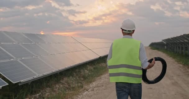 Pohled zezadu na mužského inženýra v uniformě kráčejícího ve sluneční elektrárně. Muž v těžké helmě, zkoumá předmět a nese lanovku. Koncepce rozvoje solárních stanic a zelené energie. — Stock video