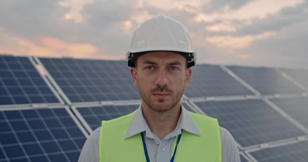 Ritratto di allegro ingegnere maschio in casco protettivo guardando alla macchina fotografica. Un bell'uomo in uniforme che sorride in piedi alla centrale solare. Concetto di sviluppo di una stazione solare. — Video Stock