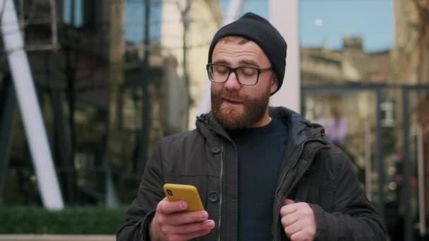 Vista de cerca de hombre barbudo en los años 30 mirando la pantalla del teléfono y sonriendo mientras camina por la calle. Chico guapo en gafas charlando en las redes sociales mientras usa el teléfono inteligente. — Vídeos de Stock